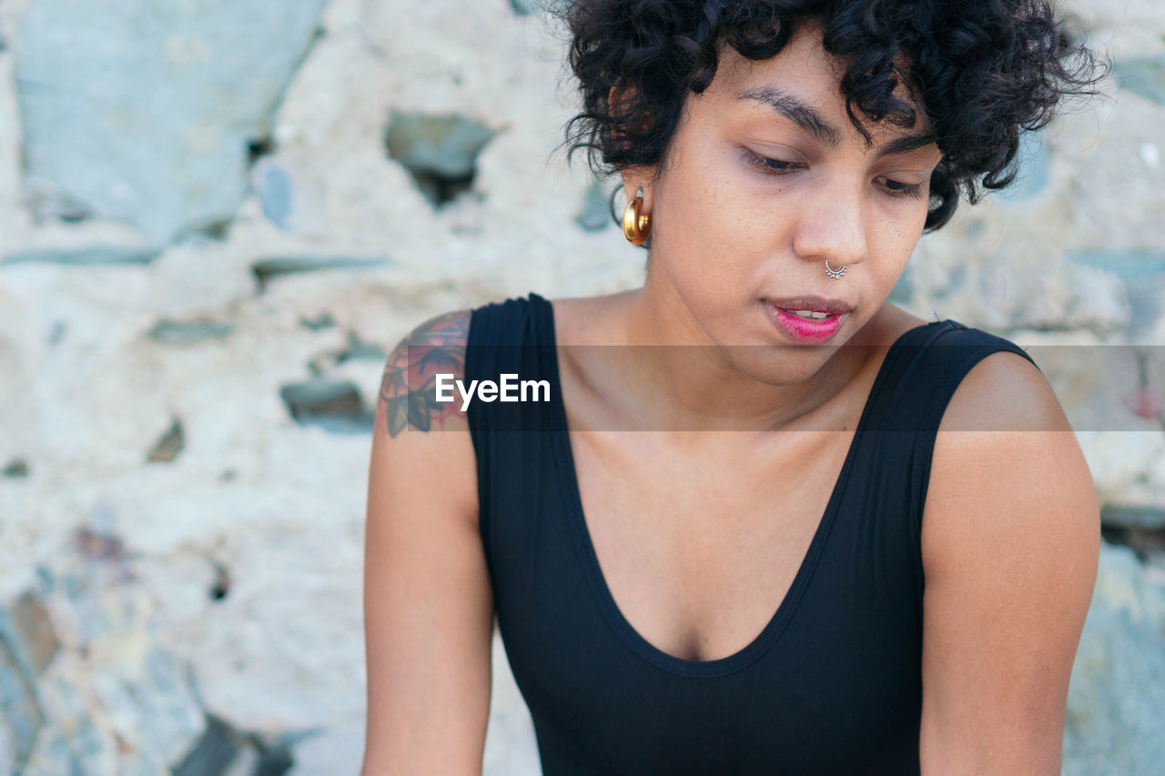 Crop melancholic young african american female with curly hair and tattoo on shoulder looking down on blurred background