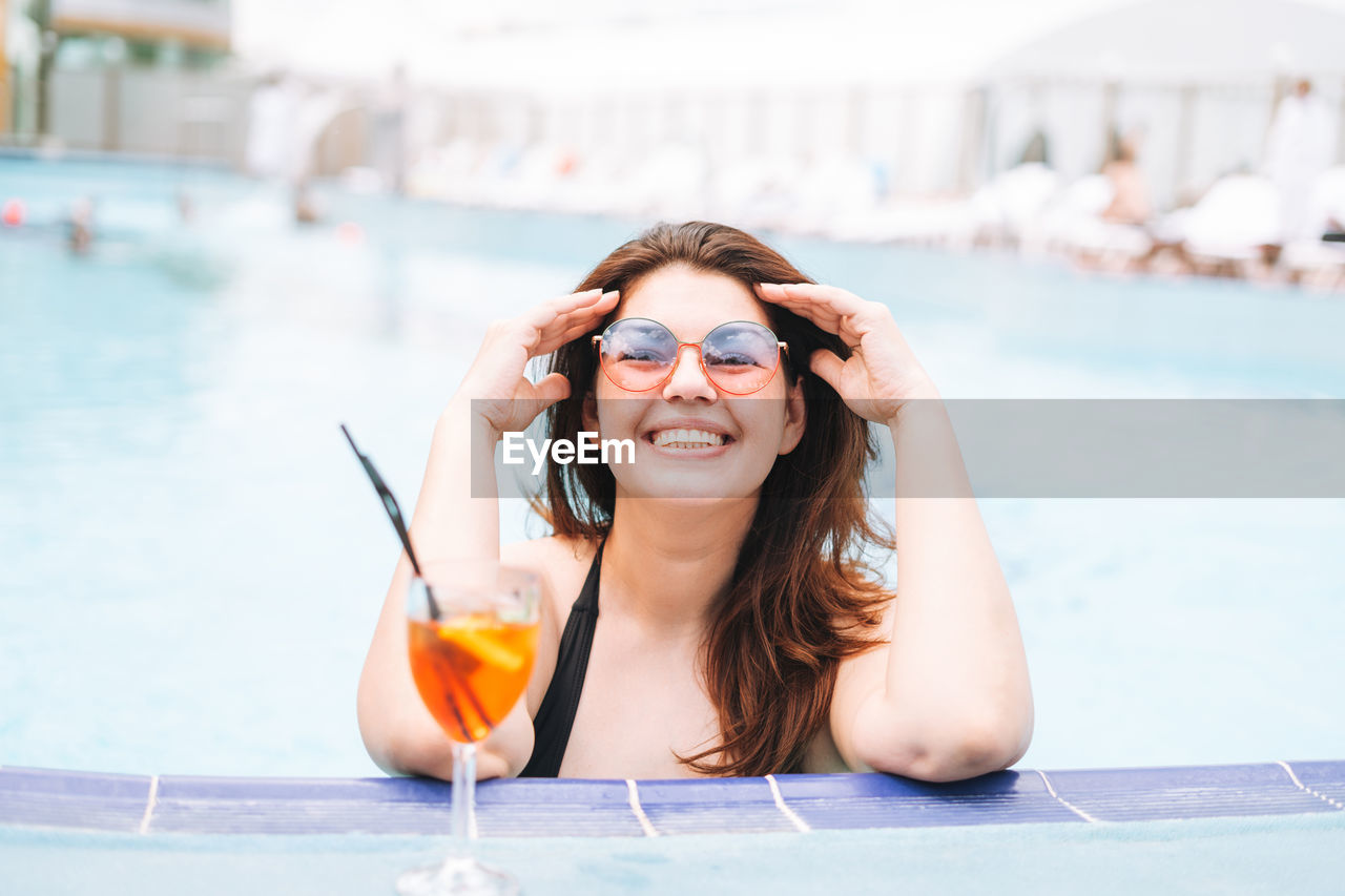 Stylish woman plus size body positive in swimsuit and sunglasses with glass of cocktail in pool