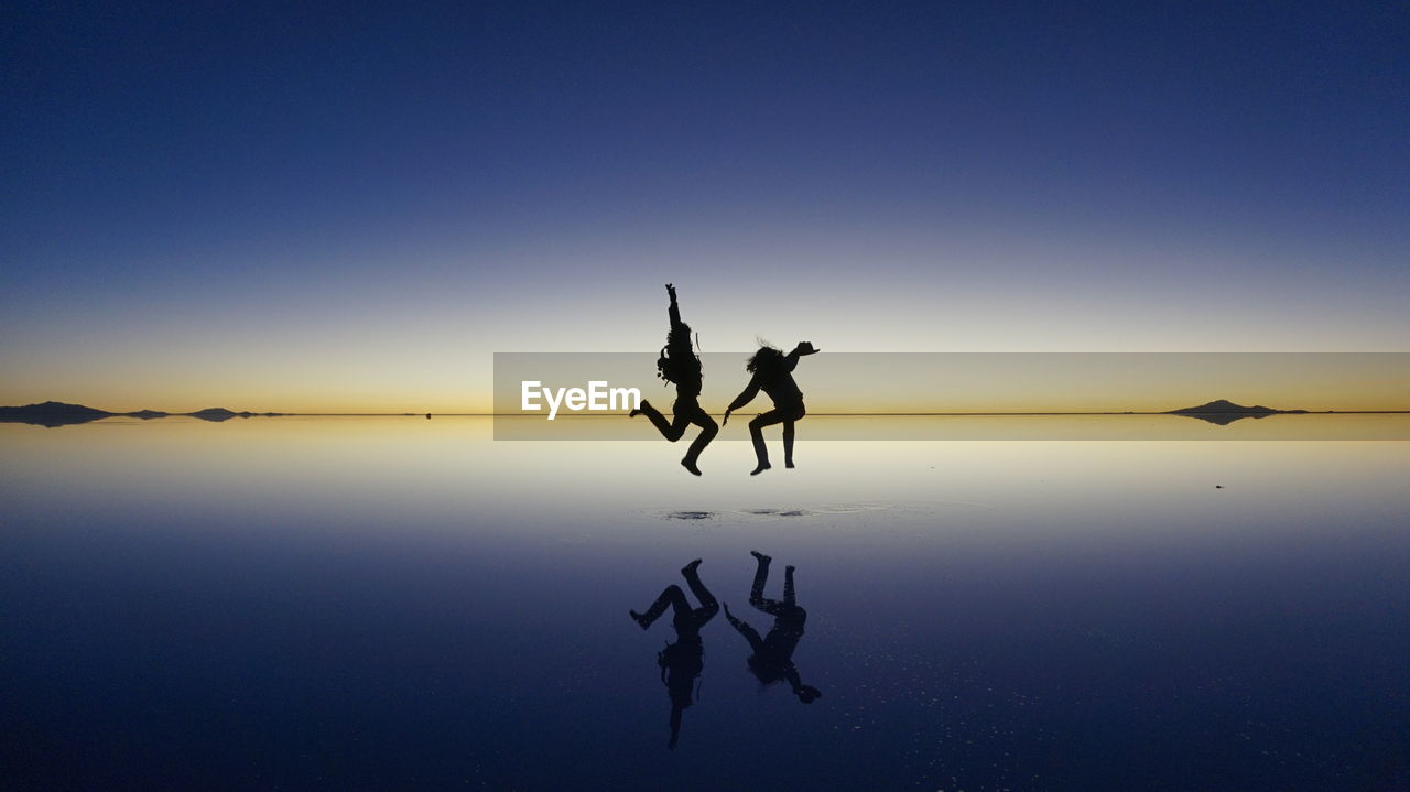 Silhouette people jumping at lake against sky during sunset