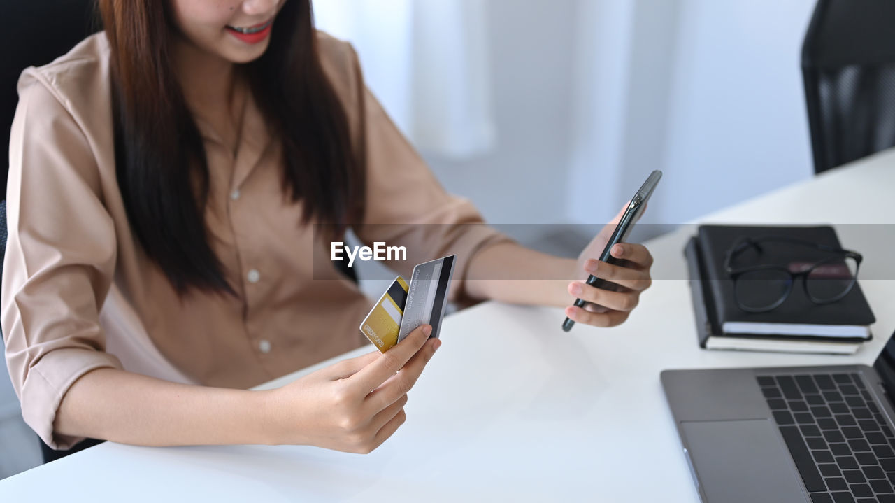 wireless technology, adult, technology, women, communication, computer, business, one person, laptop, young adult, indoors, table, portable information device, smartphone, businesswoman, internet, writing, business finance and industry, furniture, using laptop, mobile phone, working, portability, computer network, desk, long hair, convenience, person, female, office, smiling, sitting, looking, holding, occupation, happiness, clothing, lifestyles, finance, hairstyle, digital tablet, telephone, credit card, casual clothing, cheerful, white-collar worker, businesswear, looking down, buying, typing, retail, brown hair, customer, document, place of work, e-mail