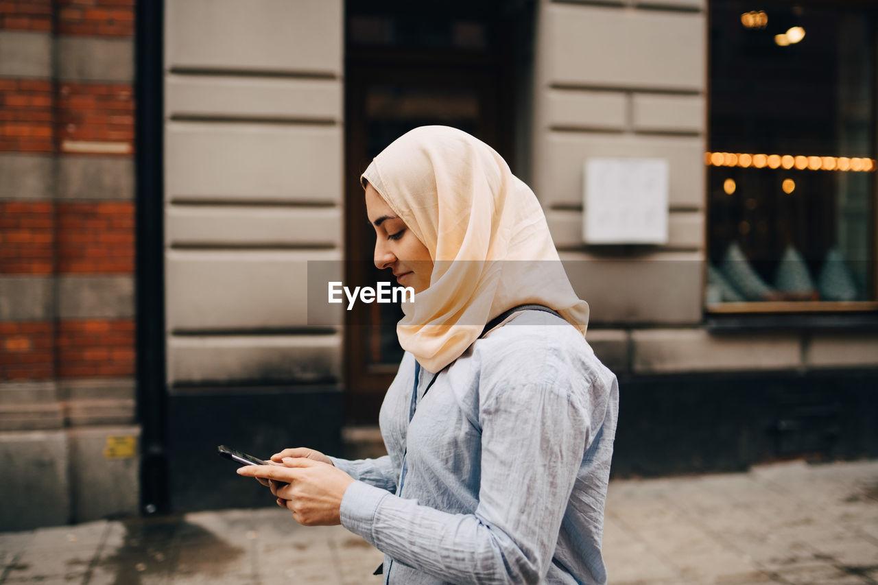 Side view of young woman using smart phone while walking on street in city