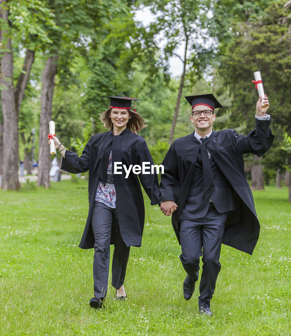 Full length portrait of couple wearing graduation gowns holding hands on field