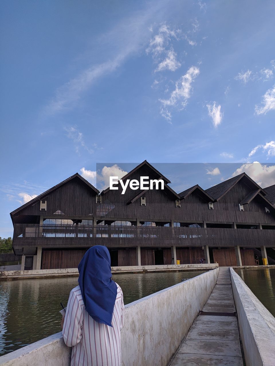 REAR VIEW OF WOMAN SITTING BY BUILDING AGAINST SKY