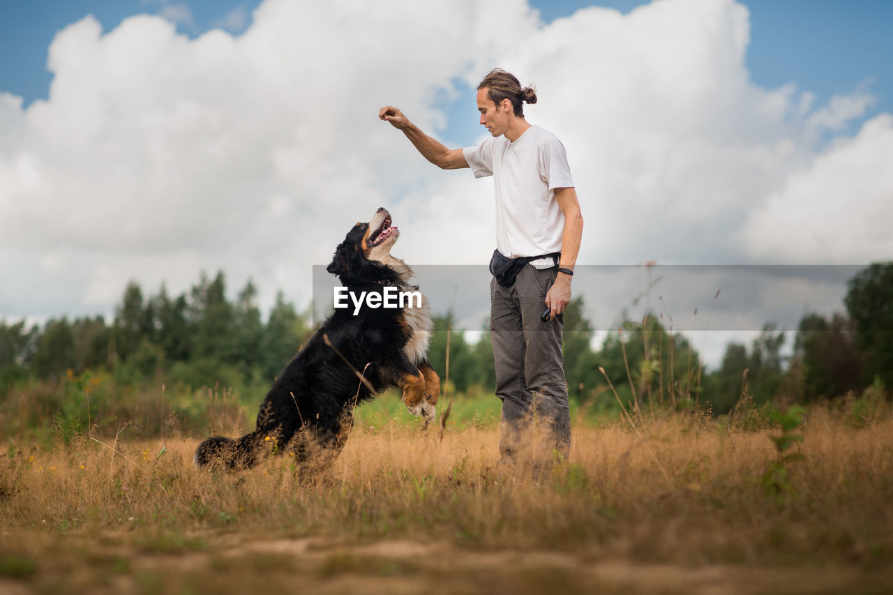 Man playing with dog while standing on grassy land against cloudy sky