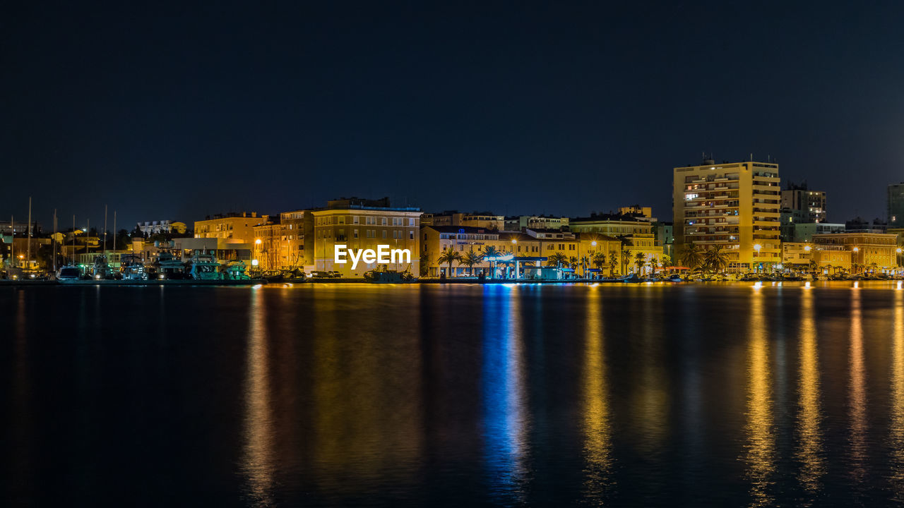 Illuminated buildings by sea against sky at night