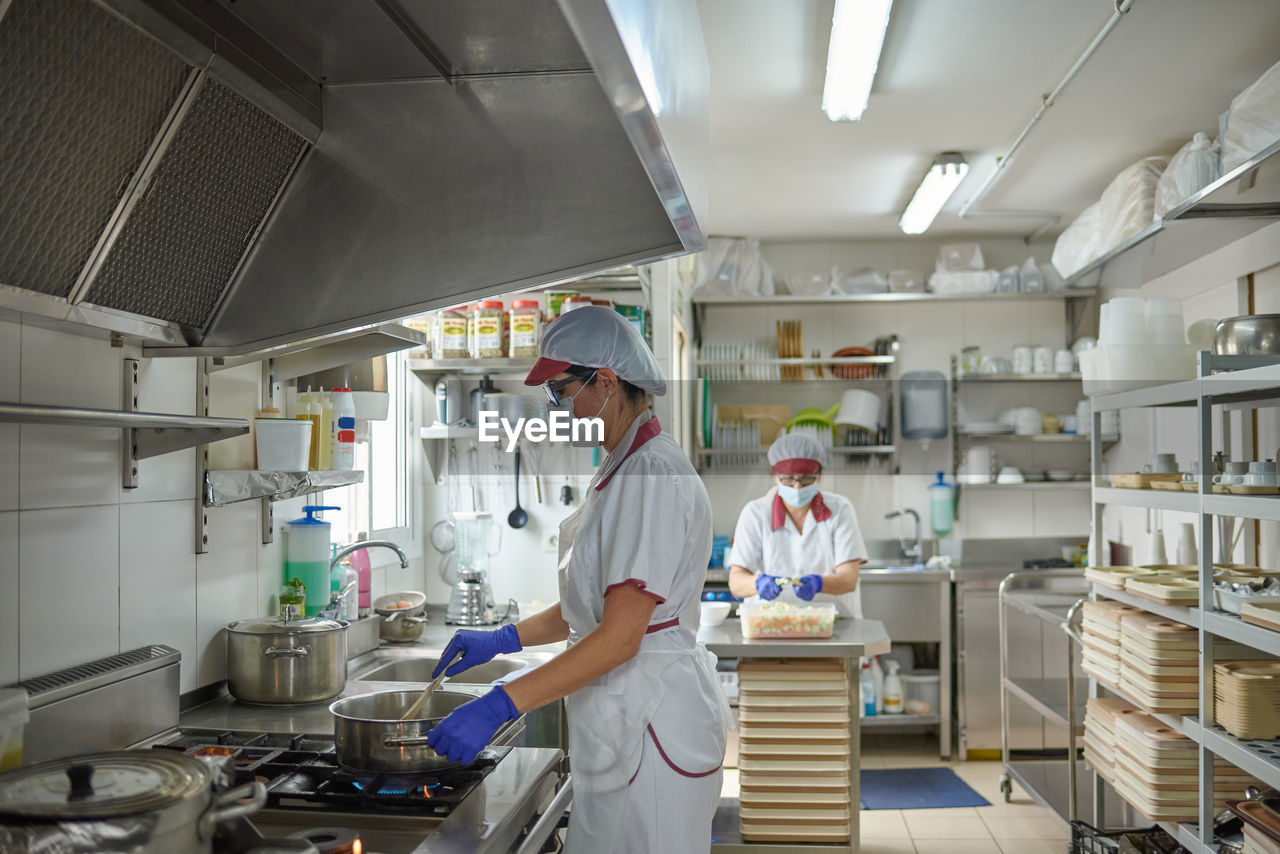 Professional female cook in white uniform and protective mask and gloves preparing food on stove while working with colleague in hospital kitchen during coronavirus pandemic