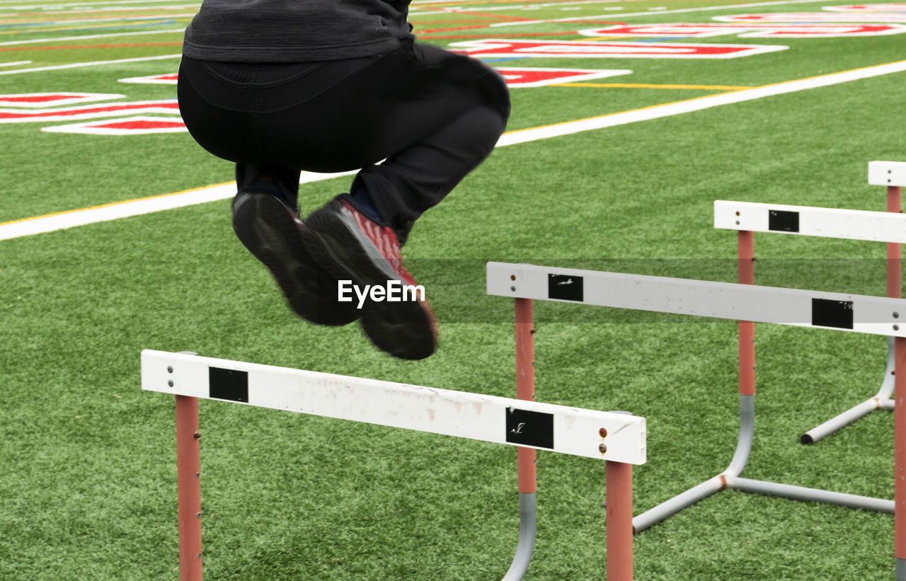 A high school boy is jumping over track hurdles on a turf field wearing sweatpants.