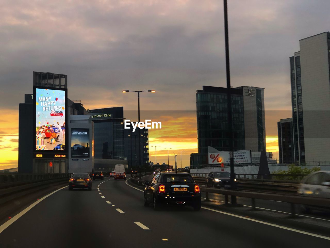 CARS ON ROAD BY BUILDINGS IN CITY AGAINST SKY AT SUNSET