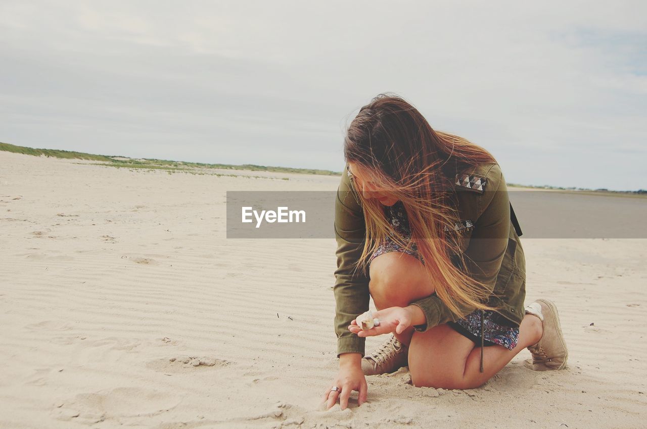 Young woman holding seashell at beach against sky