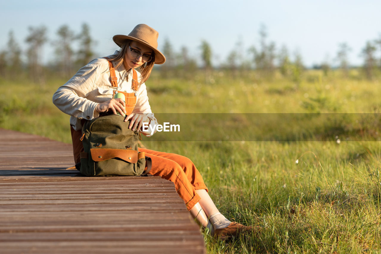 Naturalist woman in hat resting on boardwalk, takes thermos from backpack. ecotourism