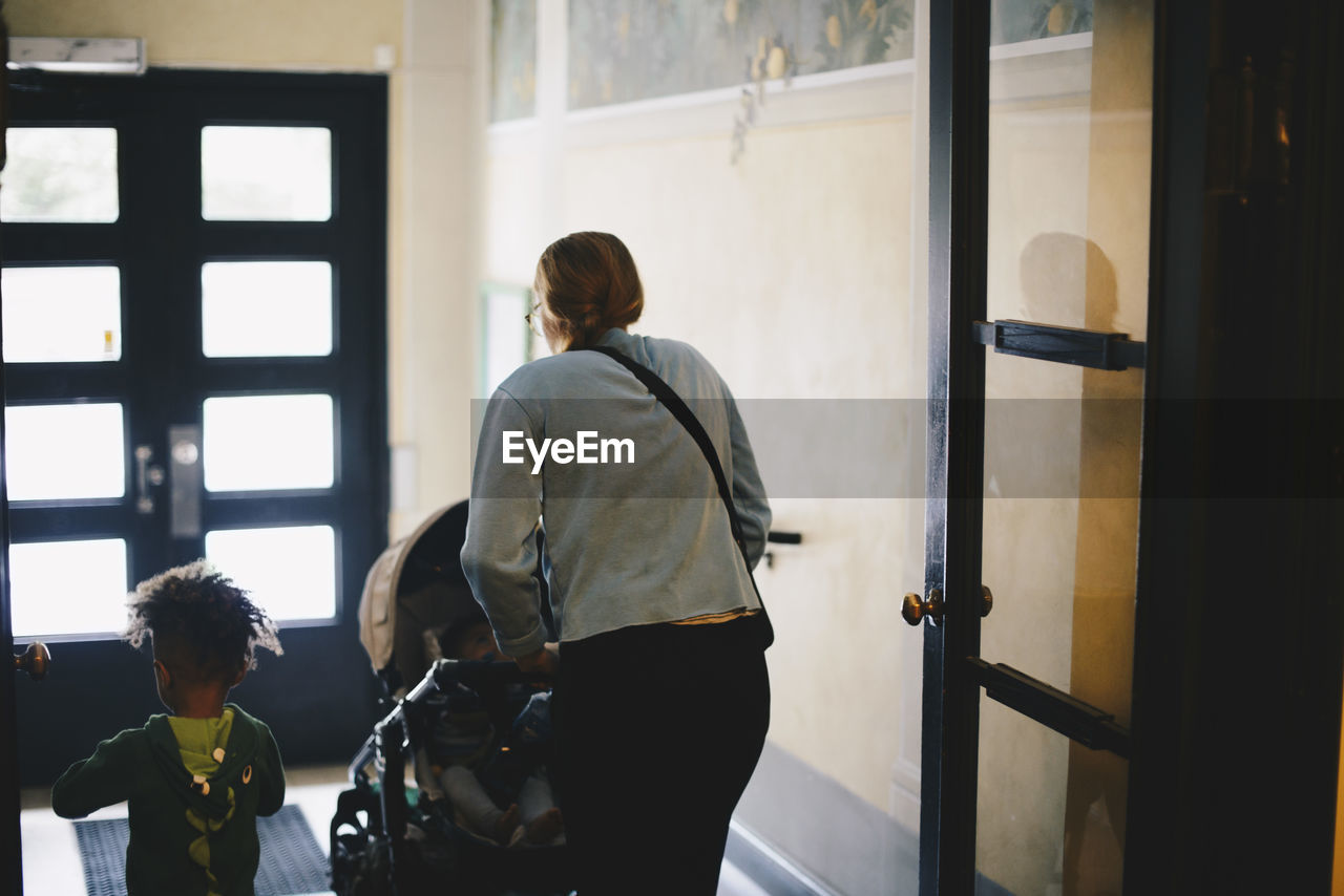 Rear view of mother pushing baby stroller while walking with son at doorway