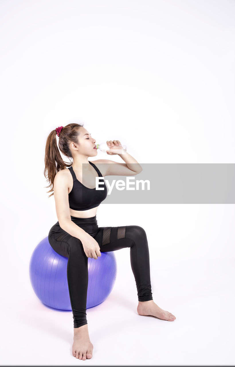 Woman drinking water while sitting on exercise ball over gray background