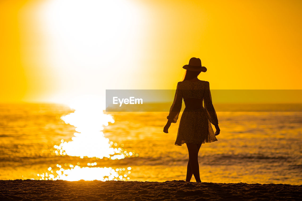 silhouette woman walking on beach against orange sky during sunset