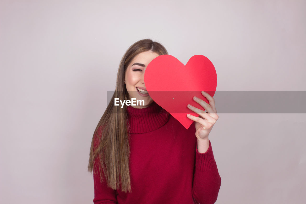 Portrait of smiling young woman holding heart shape against colored background