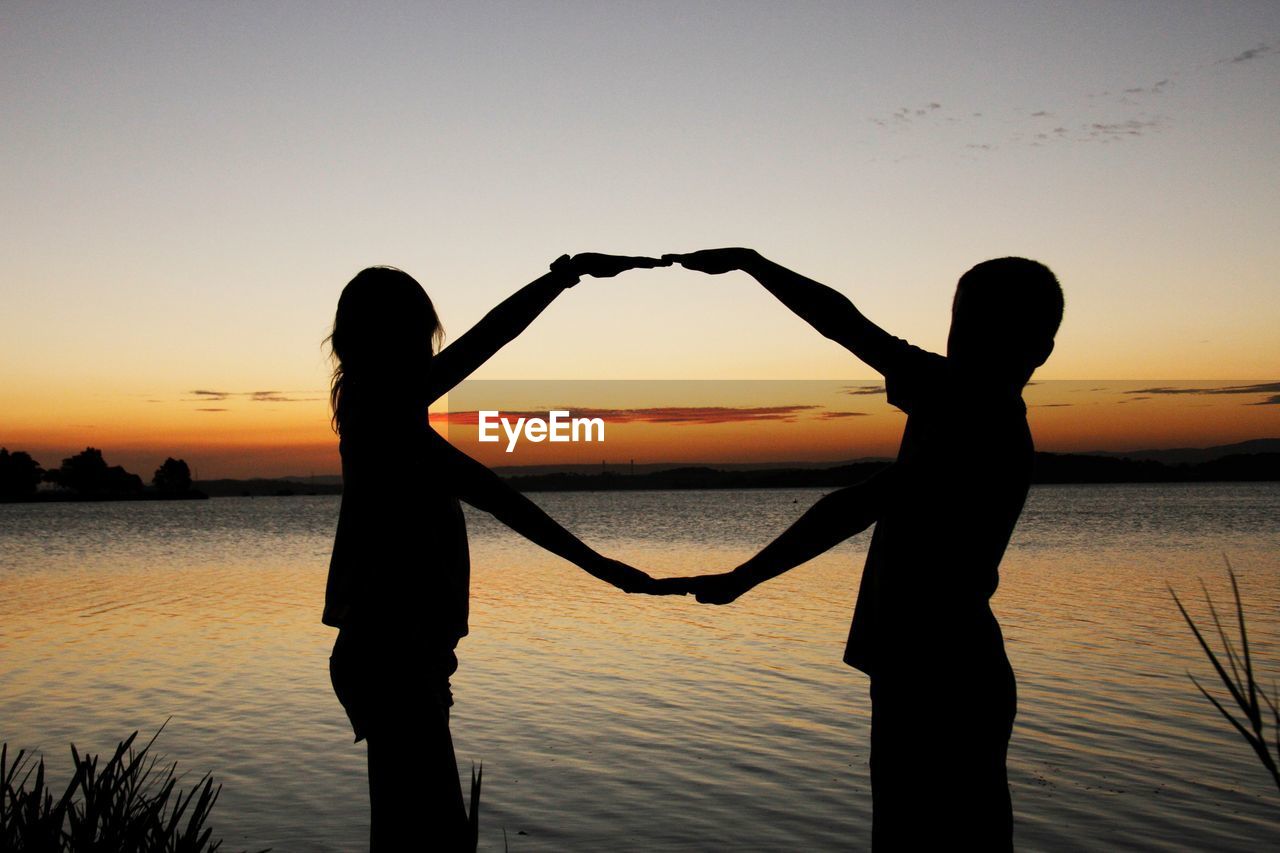 Silhouette of boy and girl making hand shape at lakeshore