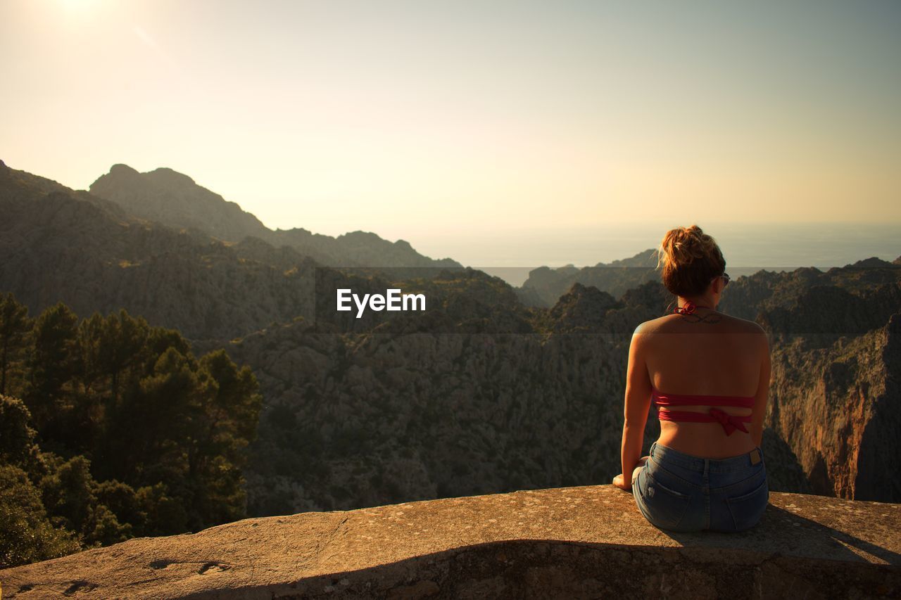 Rear view of woman sitting on retaining wall against mountains