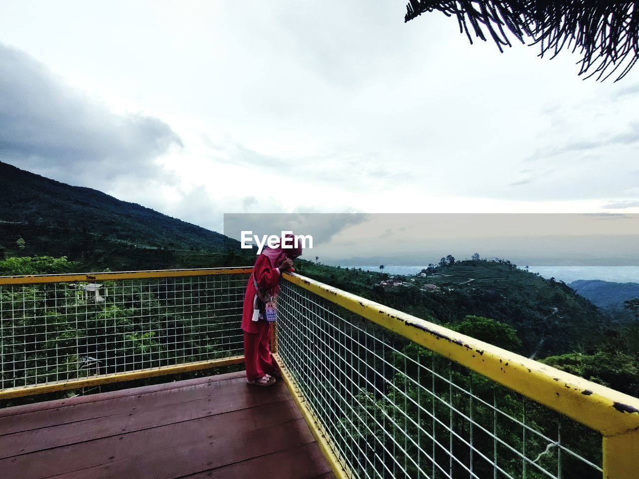 one person, railing, sky, cloud, mountain, nature, full length, adult, leisure activity, bridge, lifestyles, women, architecture, beauty in nature, sports, vacation, landscape, standing, environment, outdoors, day, clothing, scenics - nature, mountain range, travel destinations, travel, built structure, casual clothing, tranquility, activity, footbridge, walking, female, tranquil scene, rear view, holiday, young adult, transportation, plant