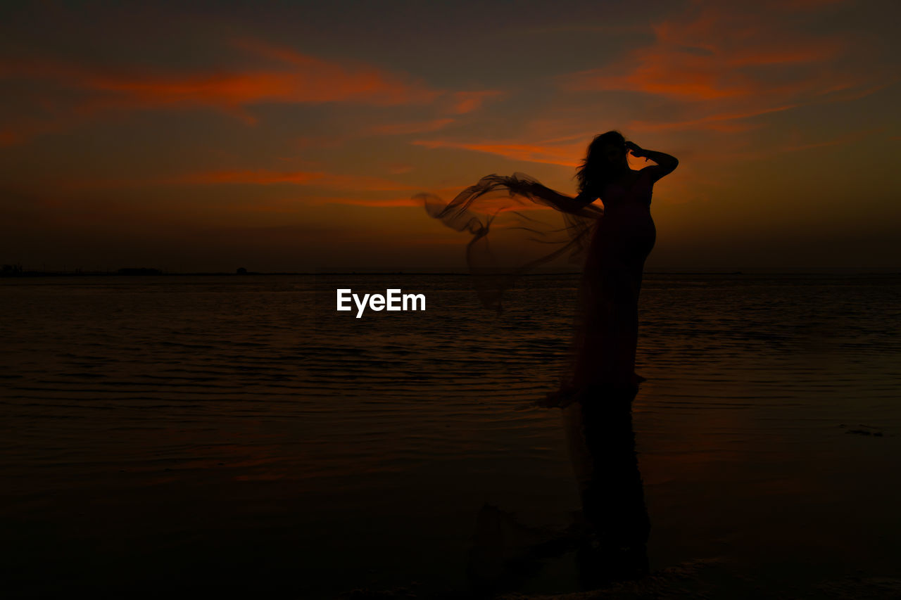 SILHOUETTE PERSON STANDING ON BEACH AGAINST ORANGE SKY