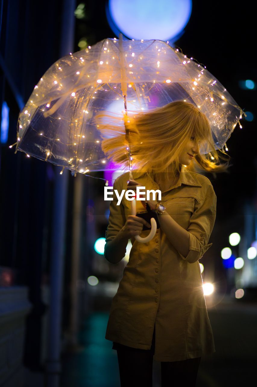 Woman tossing hair while standing with illuminated umbrella in city at night