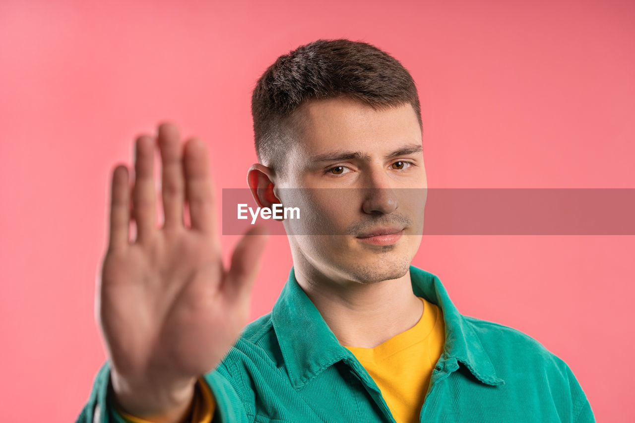 portrait, one person, studio shot, colored background, young adult, headshot, men, adult, indoors, person, looking at camera, hand, emotion, sign language, human face, finger, front view, gesturing, serious, casual clothing, looking, buzz cut, clothing, copy space, human head