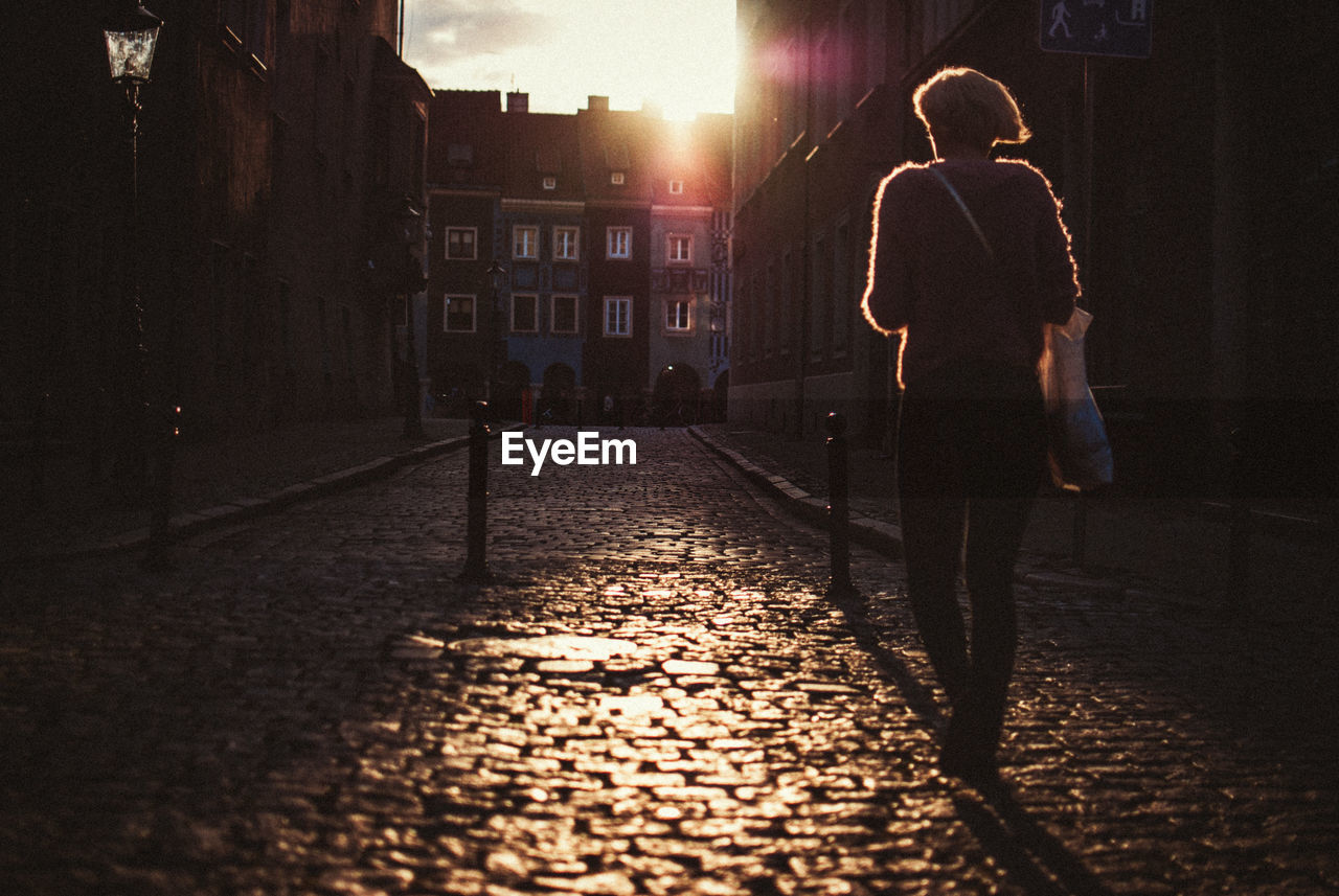 Rear view of woman walking on street amidst buildings during sunset