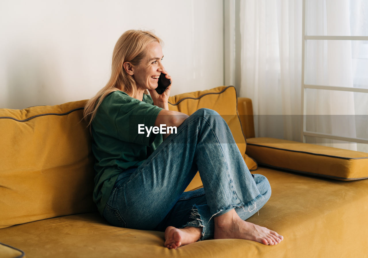 Attractive middle aged woman chatting cheerfully on the phone while sitting on the couch. person