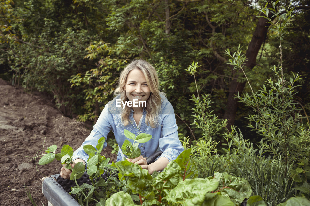 PORTRAIT OF A SMILING YOUNG WOMAN HOLDING PLANTS IN FOREST