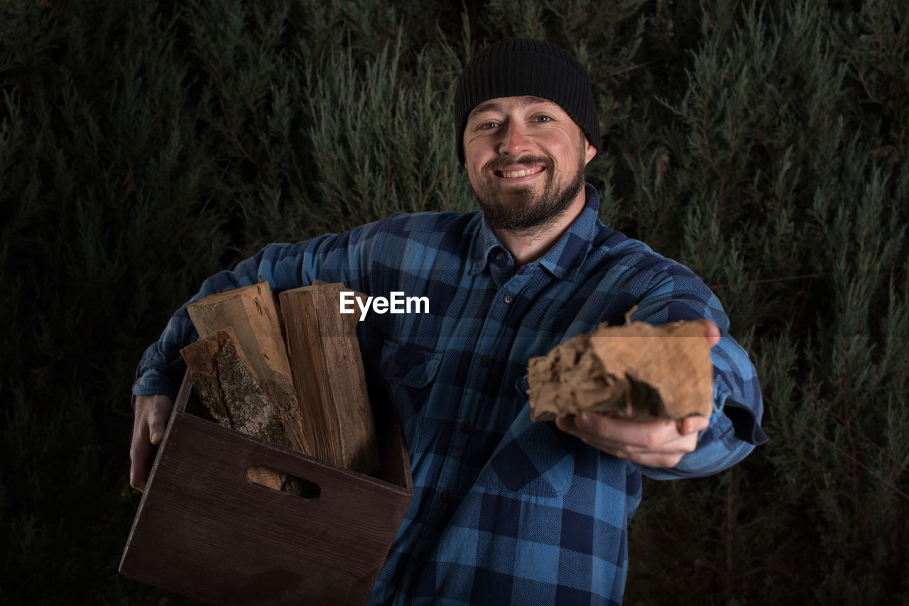 Portrait of smiling lumberjack holding wood and crate against trees at forest