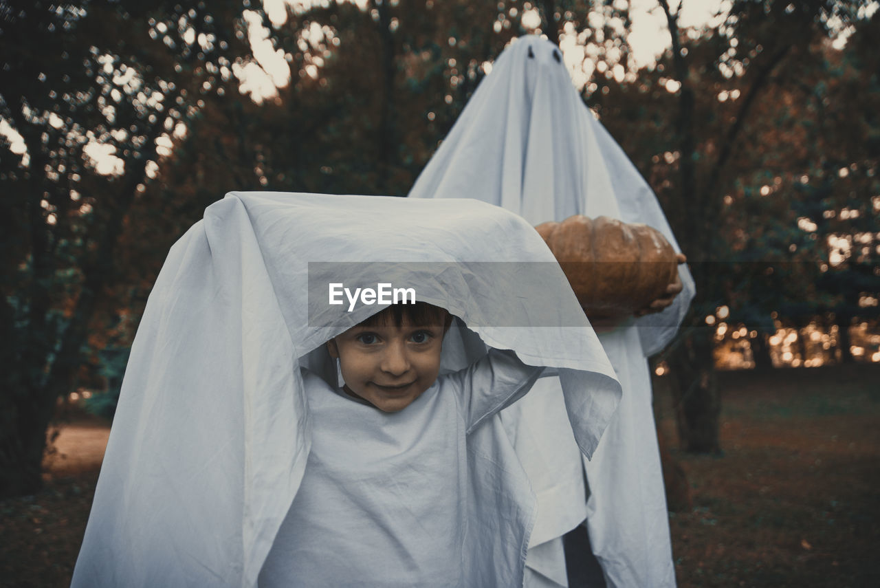 Portrait of cute boy with person wearing ghost costume in park at sunset during halloween