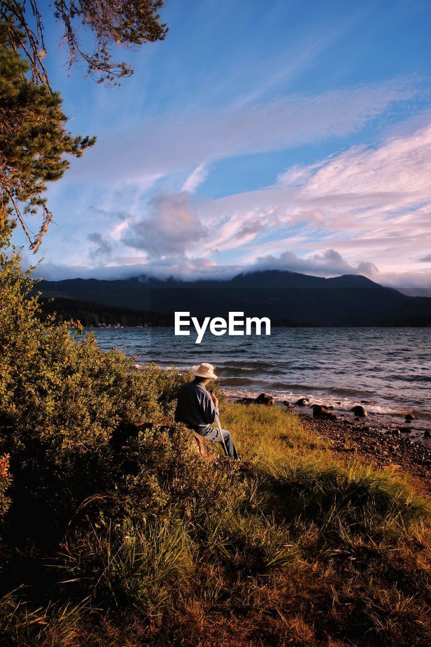 Rear view of senior man sitting by lakeshore during sunset