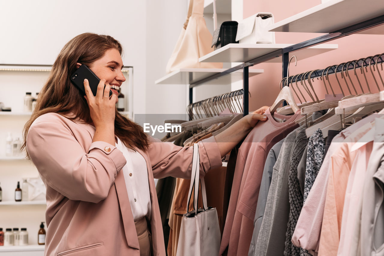 Smiling woman talking on cell phone while choosing clothes at rack in fashion store	