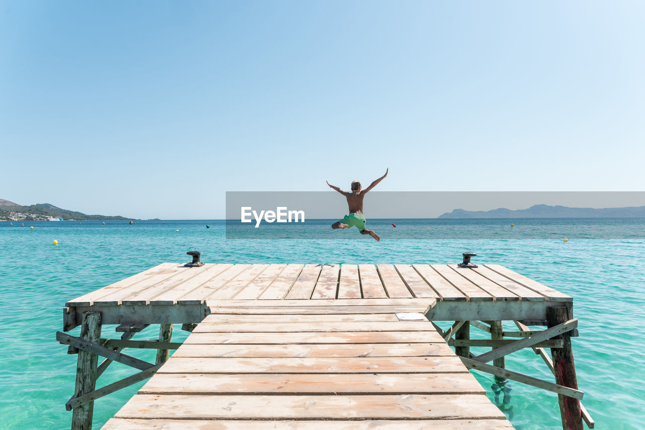 Back view of unrecognizable teenage boy in moment of jumping from pier in sea enjoying summer weekend on sunny day