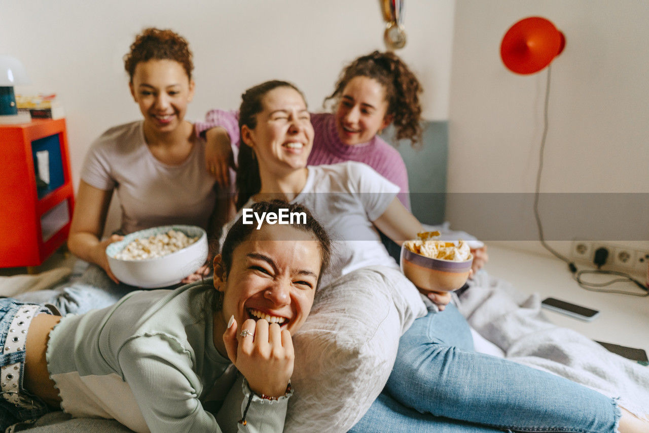 Cheerful female friends watching tv while enjoying snacks at home