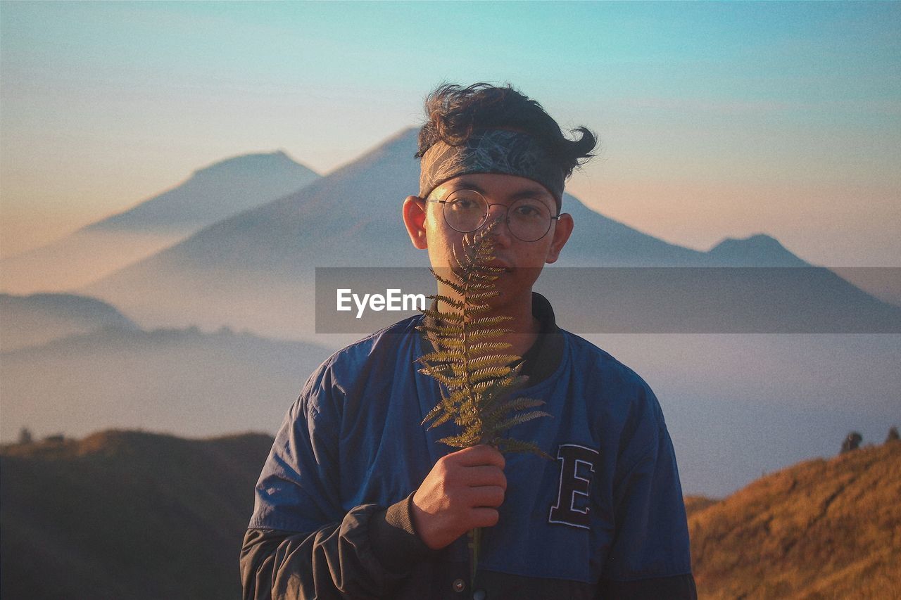 PORTRAIT OF YOUNG MAN STANDING AGAINST MOUNTAIN DURING SUNSET