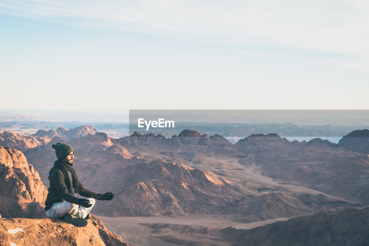 MAN SITTING ON ROCKS AGAINST MOUNTAIN