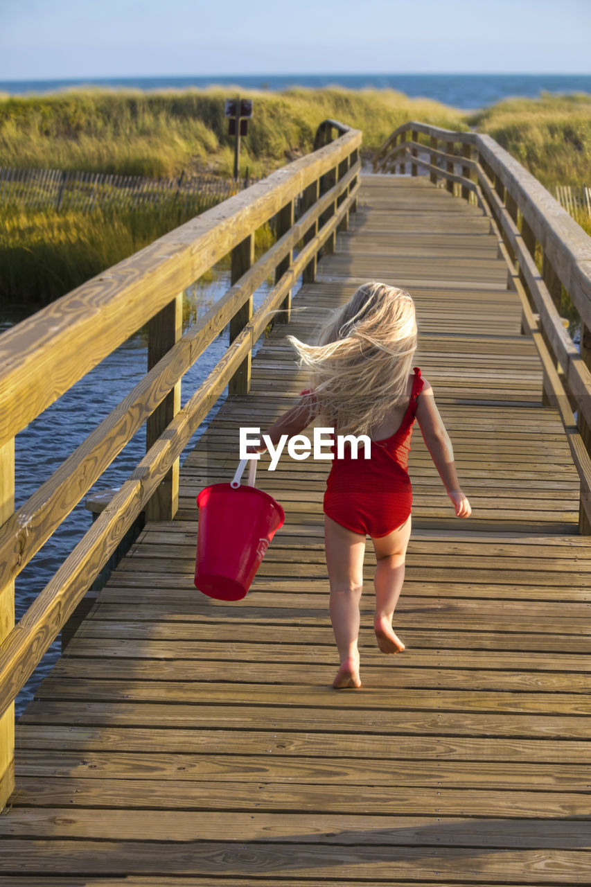 Little girl from behind running on bridge to beach with red bucket