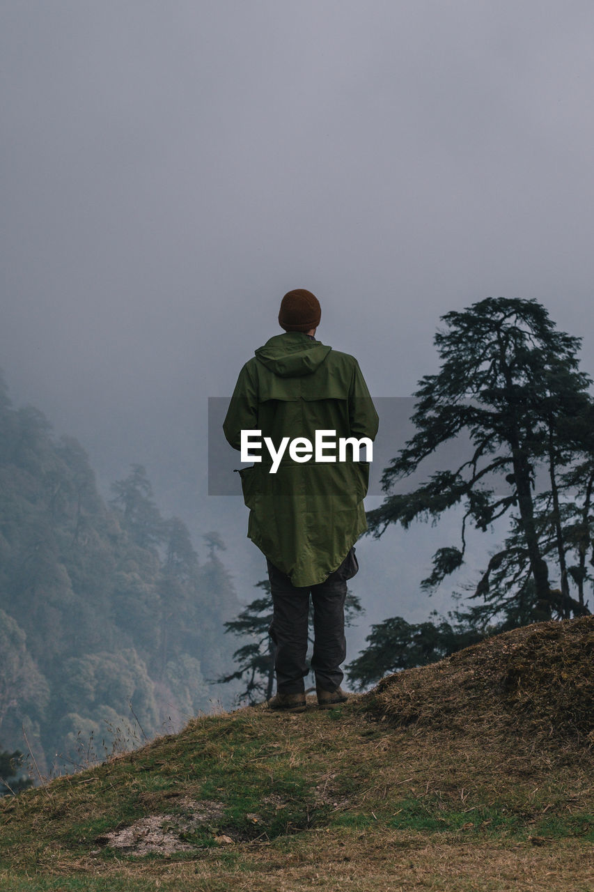 REAR VIEW OF MAN STANDING BY TREE ON MOUNTAIN