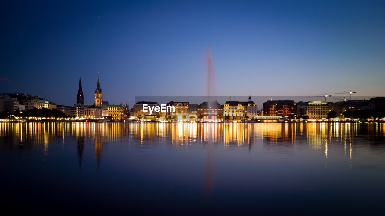 Illuminated buildings by river against sky at dusk