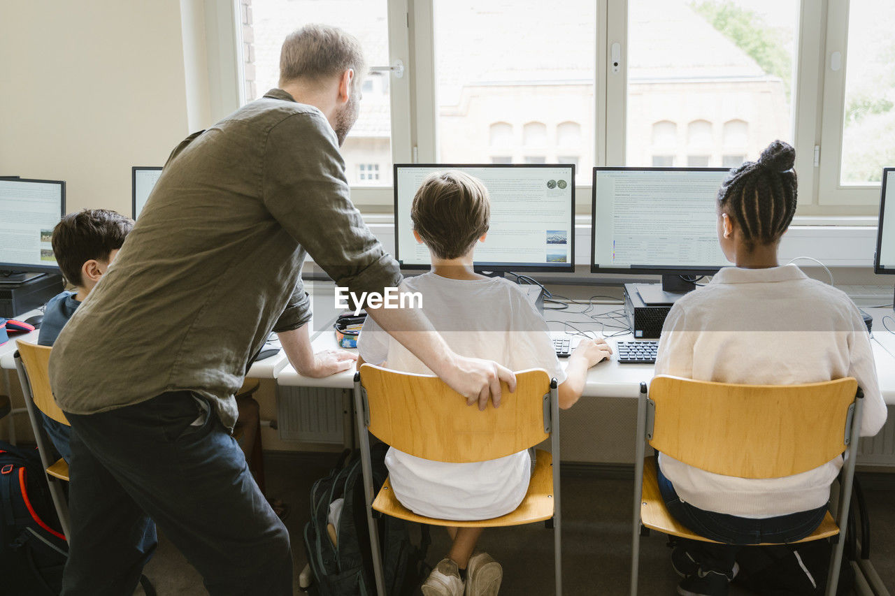 Male teacher assisting students surfing internet through computers in classroom at school