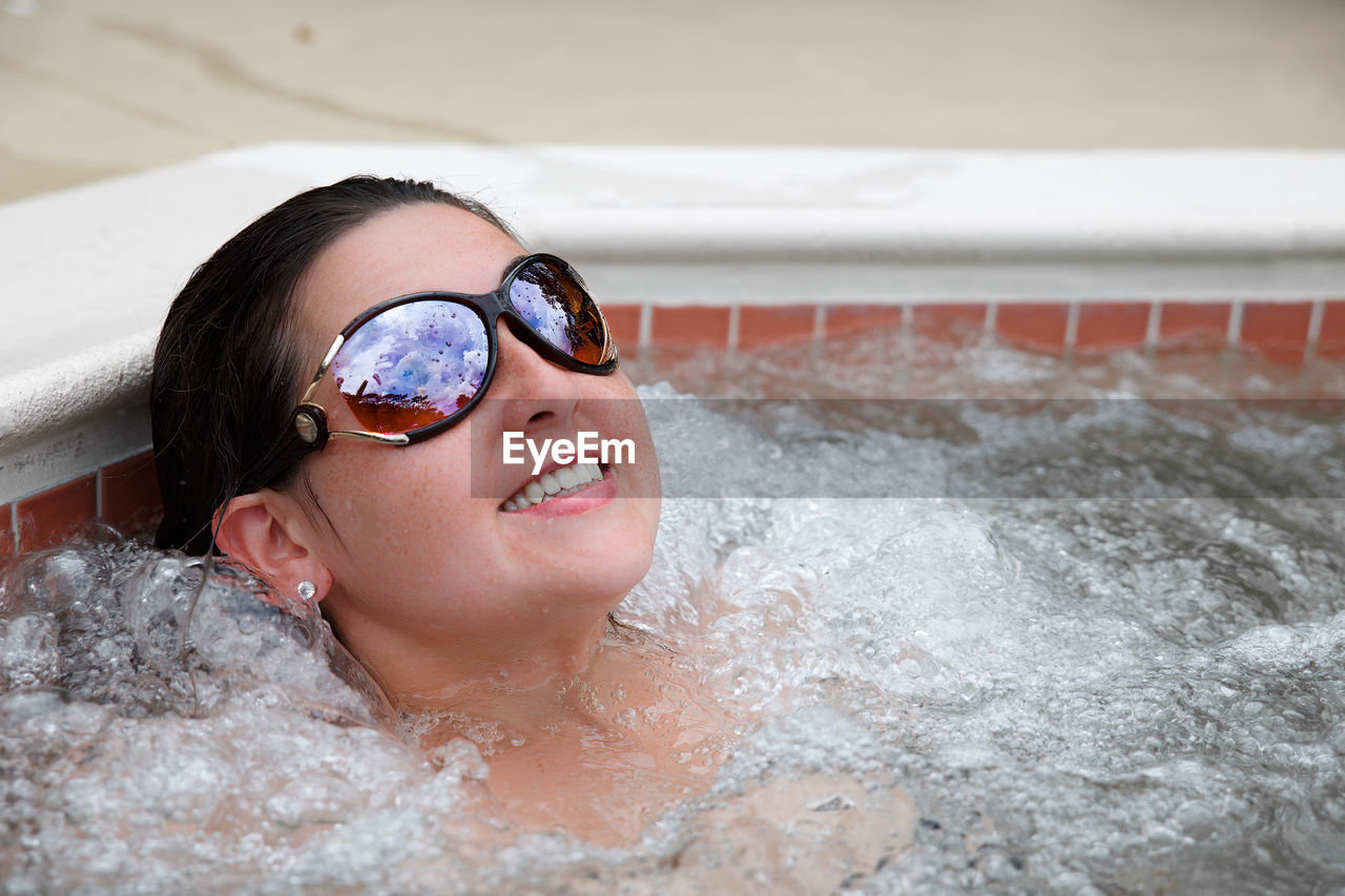 Young adult woman brunette lounging in hot tub with sunglasses smiling with bubbles