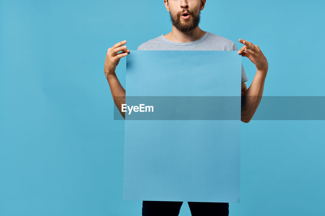 one person, studio shot, colored background, blue, adult, blue background, standing, indoors, men, young adult, t-shirt, front view, copy space, beard, portrait, facial hair, clothing, casual clothing, sleeve, lifestyles, person, three quarter length, looking, looking at camera, emotion, waist up, exercising, sports clothing, holding, athlete