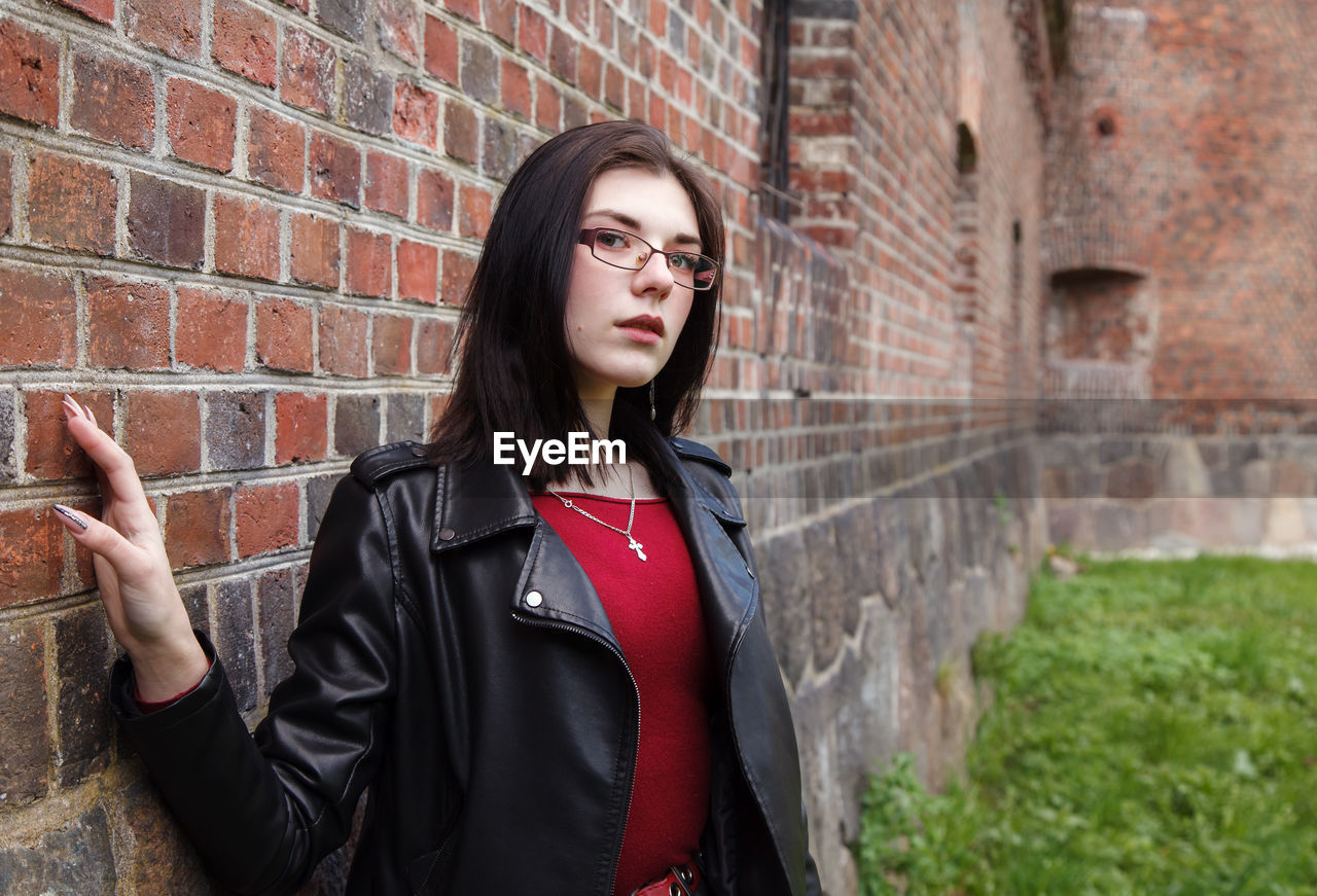 Young beautiful girl in black jacket and jeans stands near the old fortress wall on sunny day