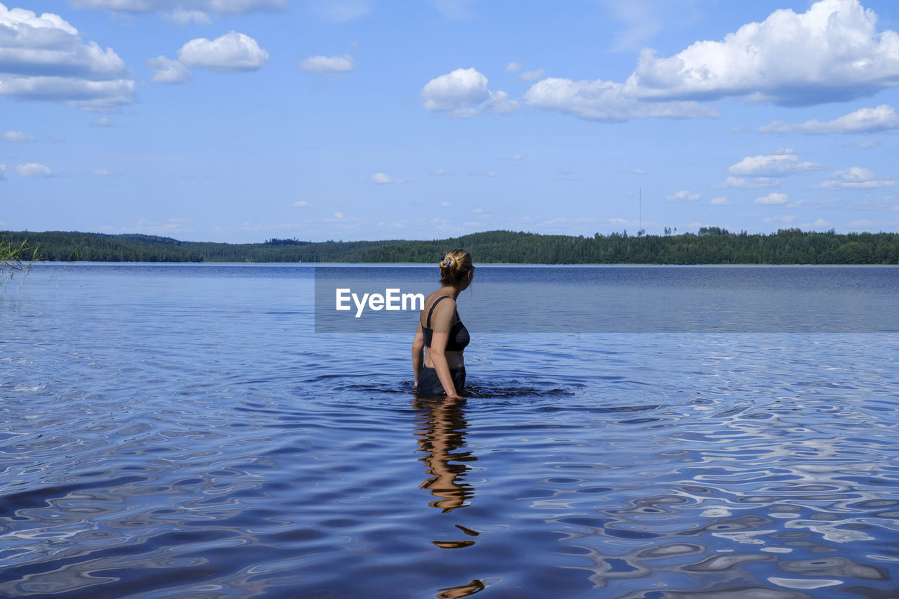 REAR VIEW OF SHIRTLESS MAN STANDING IN LAKE AGAINST SKY