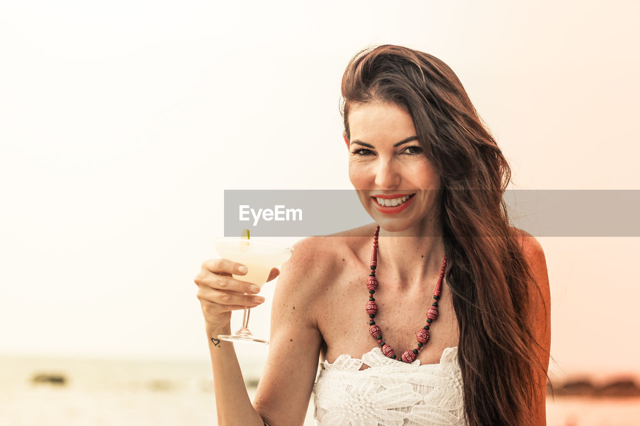 Portrait of mature woman holding drink at beach against sky