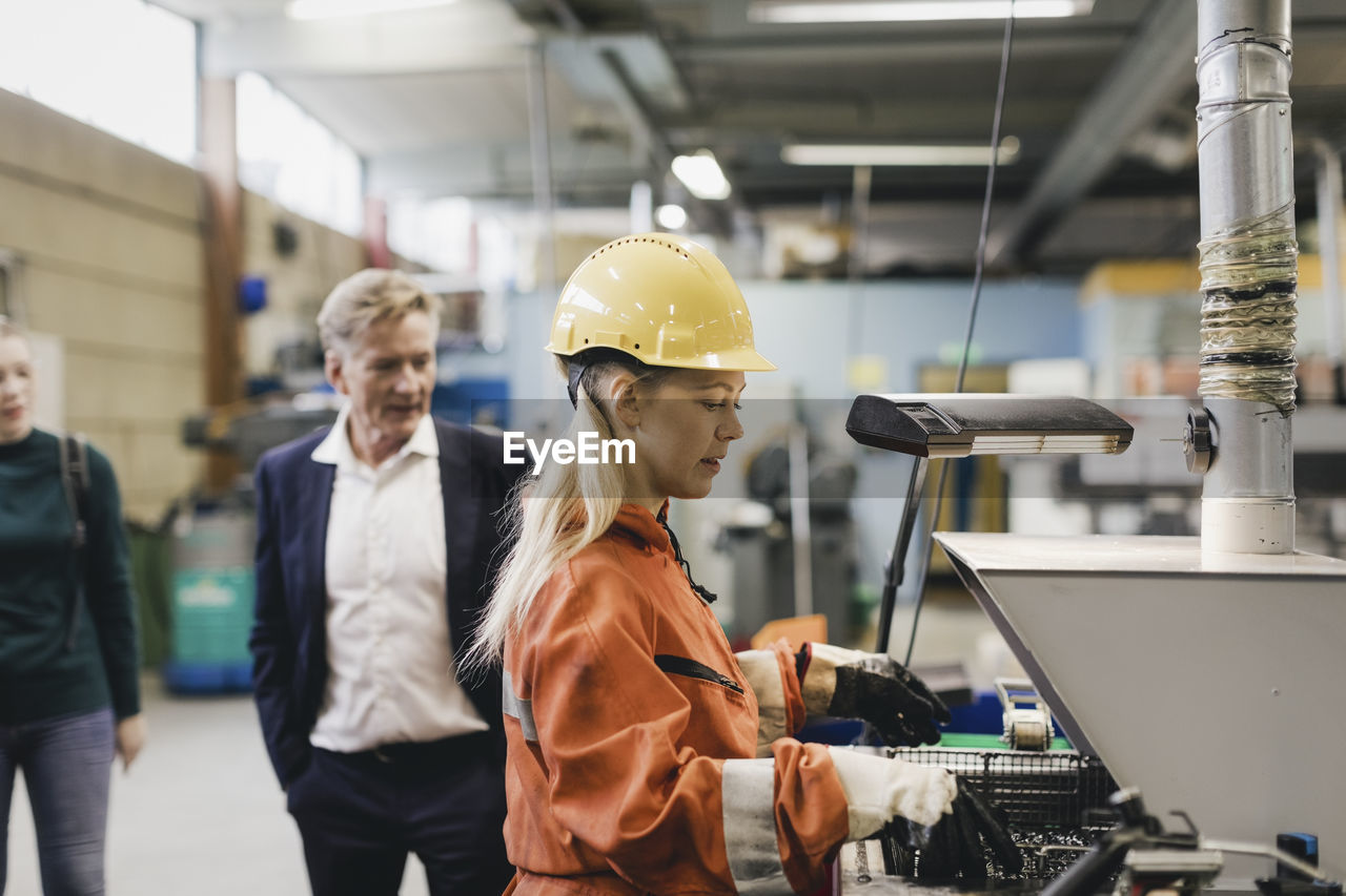 Female industrial worker in hardhat working on machinery while managers in background at factory