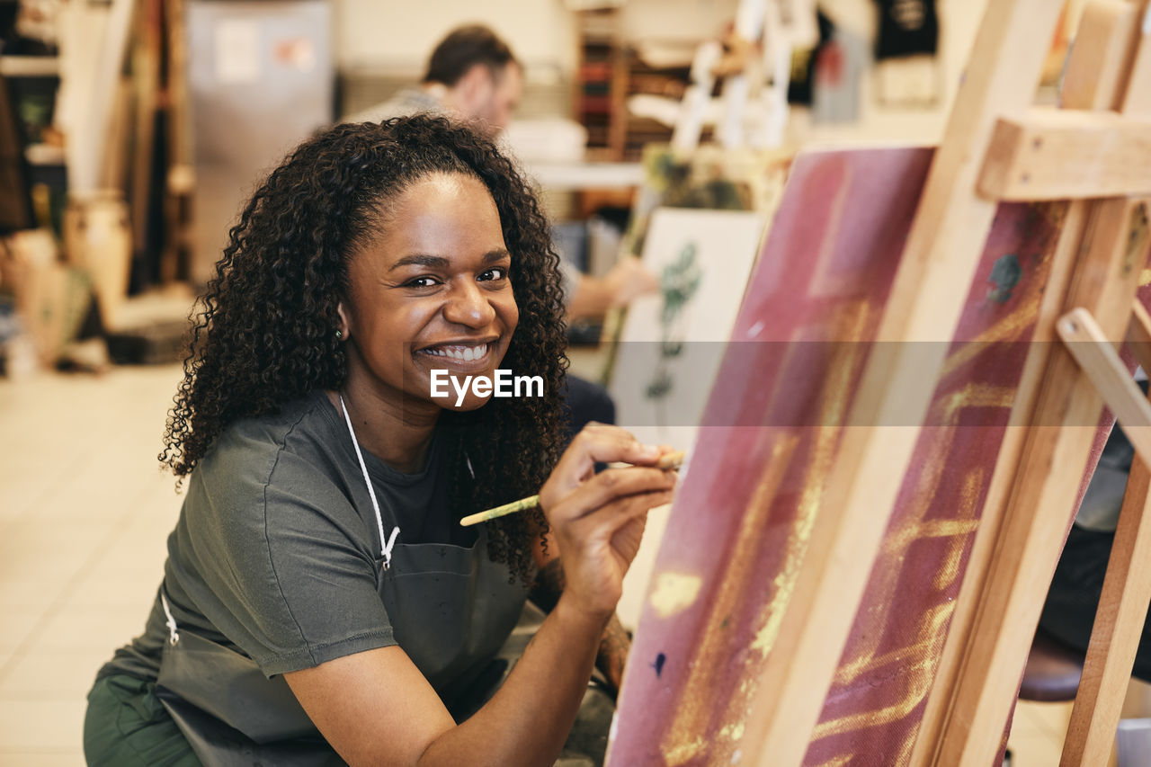 Portrait of smiling mature woman with paintbrush sitting by easel in art class