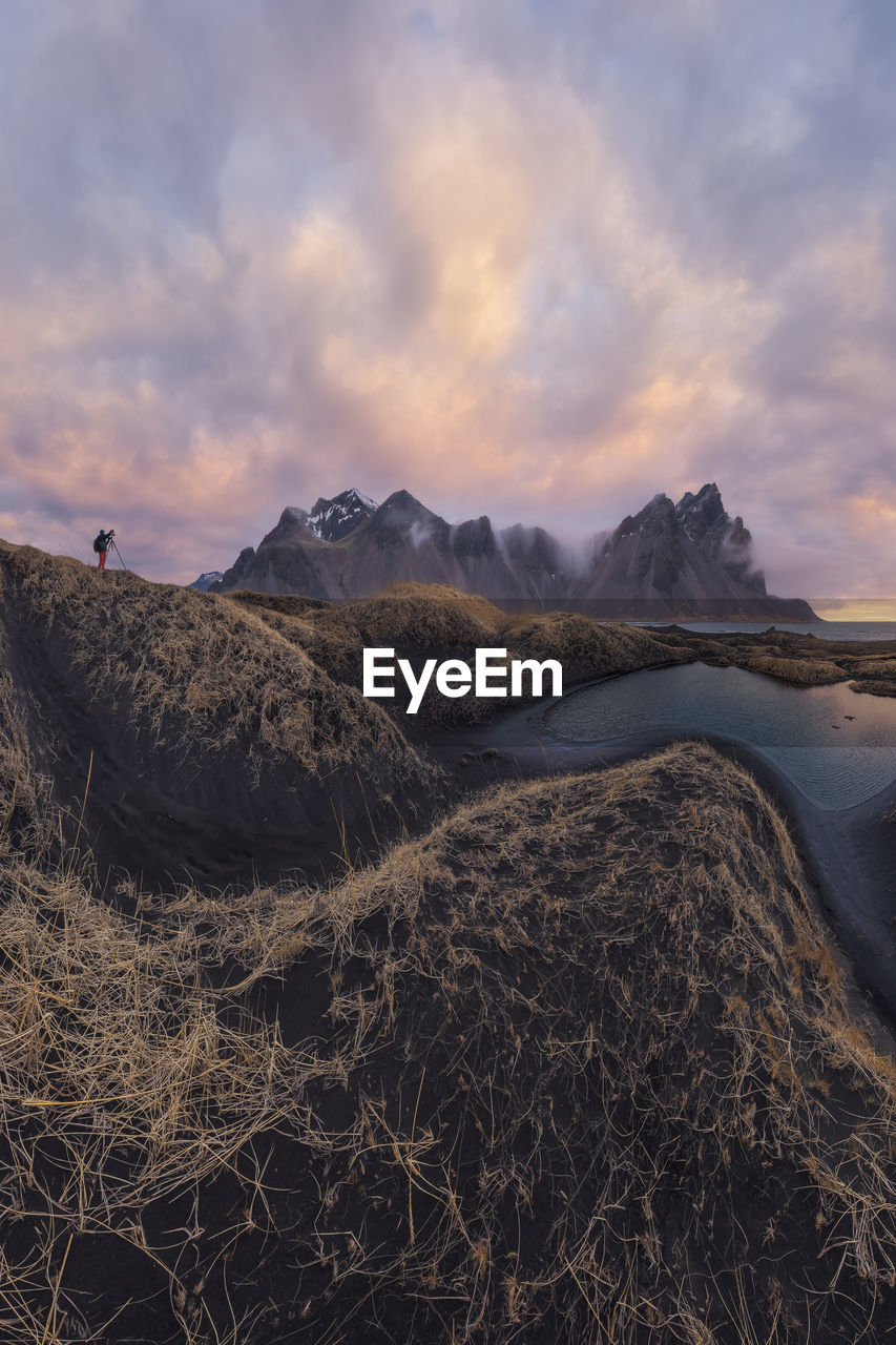 Distant traveler standing on top of mountain range against cloudy sky during sunset near black sand beach in iceland