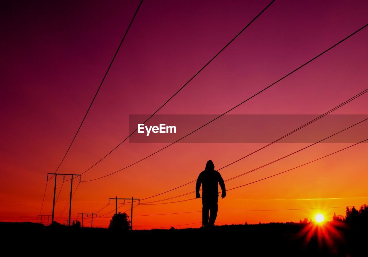 Silhouette man walking against electricity pylon during sunset