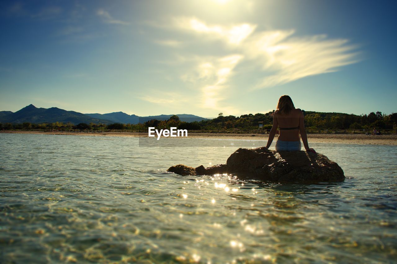 Woman sitting on rock in sea against sky during sunset