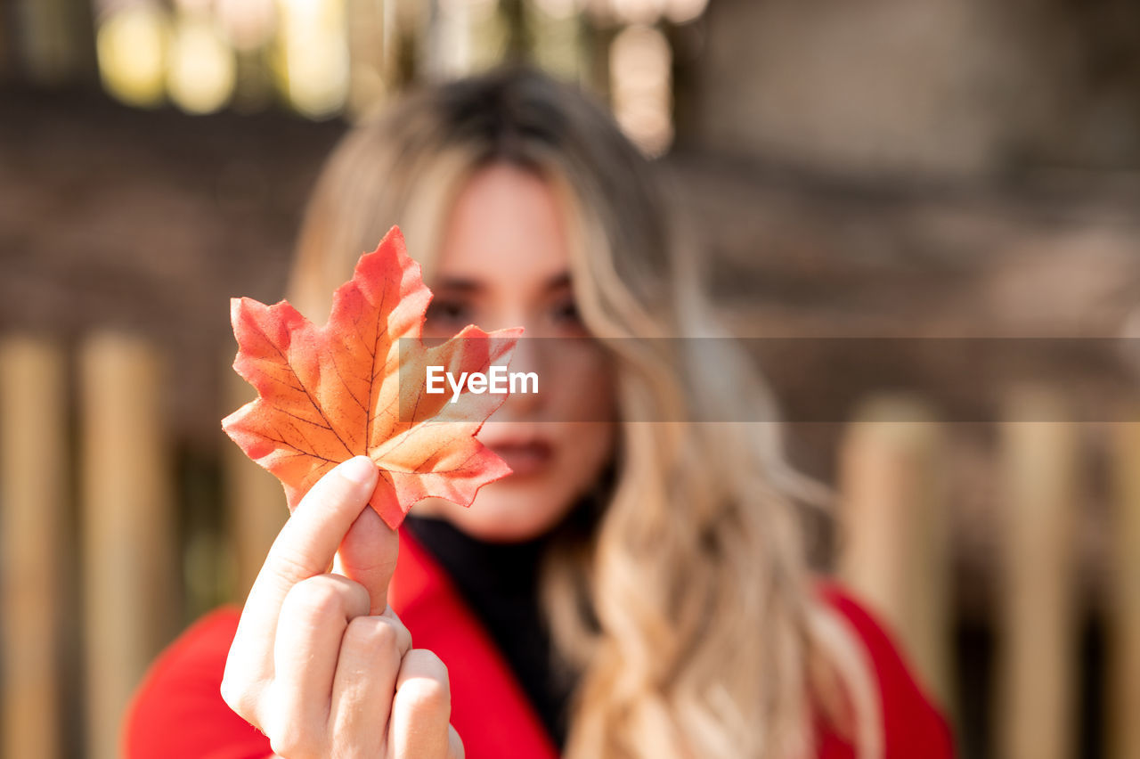 Blonde-haired woman in a red coat holding an autumn leaf of a tree with her hand in a park